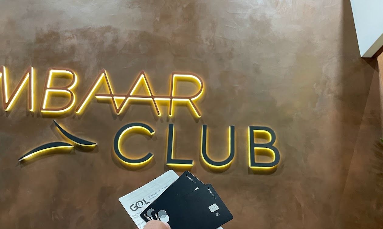 Dois cartões C6 Carbon e um cartão de embarque em frente ao letreiro da sala VIP Ambaar Club